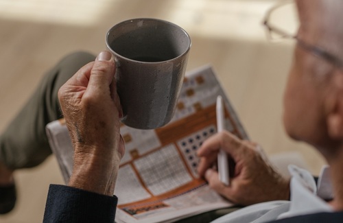 Over the shoulder view of a person holding a mug in the left hand and a pen in their right and a newspaper crossword on their lap