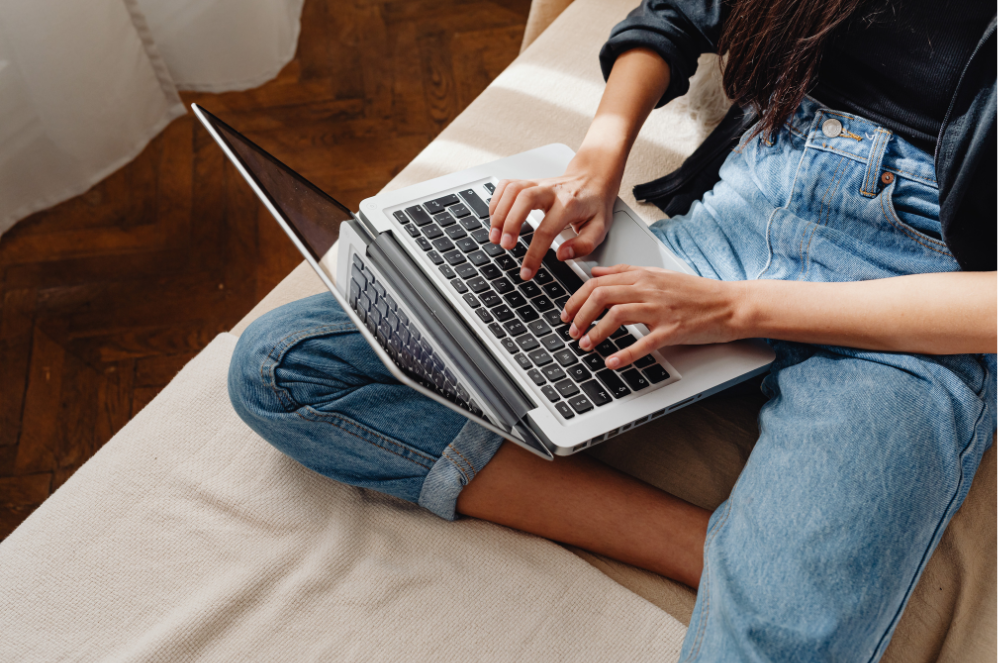 A young person sitting cross-legged on a couch and typing on their laptop