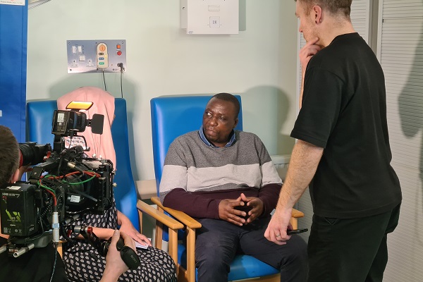 Patient sitting in a hospital chair with a crew and camera filming them for television