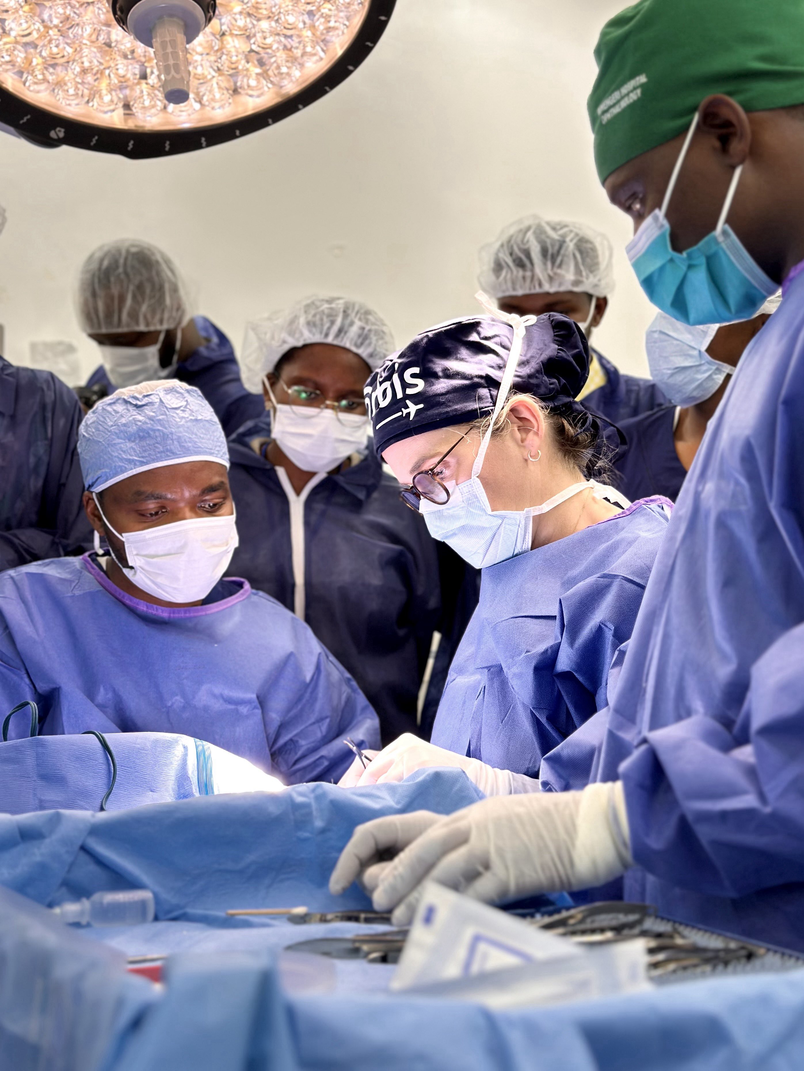 Three people performing surgery with viewers behind wearing medical PPE