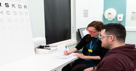 two people sitting at a desk, one person is making notes and the other is undertaking an eye examination