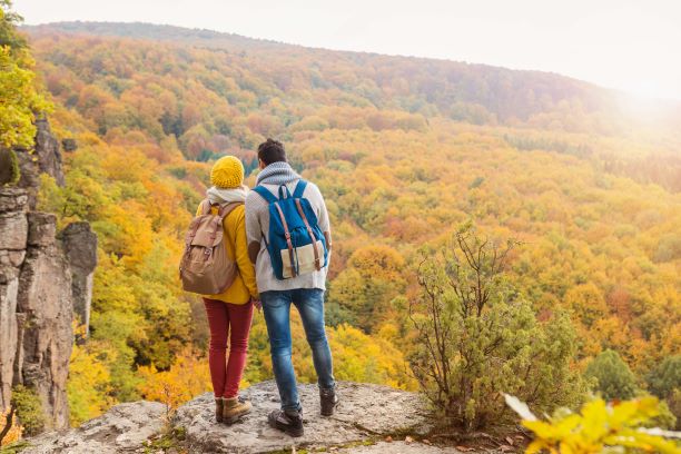 Two people with outdoor clothes, backpacks and hiking boots, standing on rocks overlooking an autumnal woods